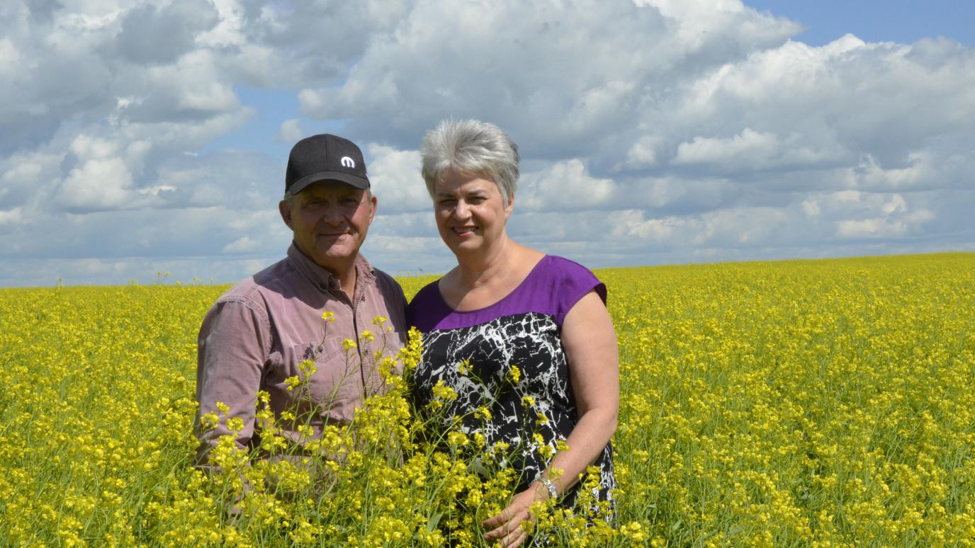 A man and a woman stand in a field with tall green grass in front of a blue sky.