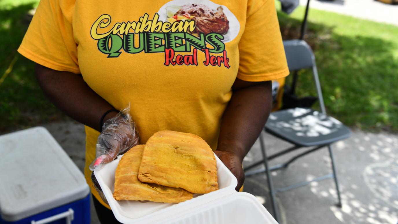 A food vendor at the 2025 Multicultural Festival holds a takeout container containing two yellow hand pies. Their shirt reads Caribbean Queens Real Jerk.