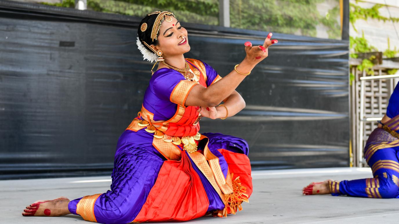 A performer at the 2025 Multicultural Festival wears a purple and red traditional outfit while giving a dance performance on stage. They are seat on the ground, and a red-painted hand stretches out towards the crowd.