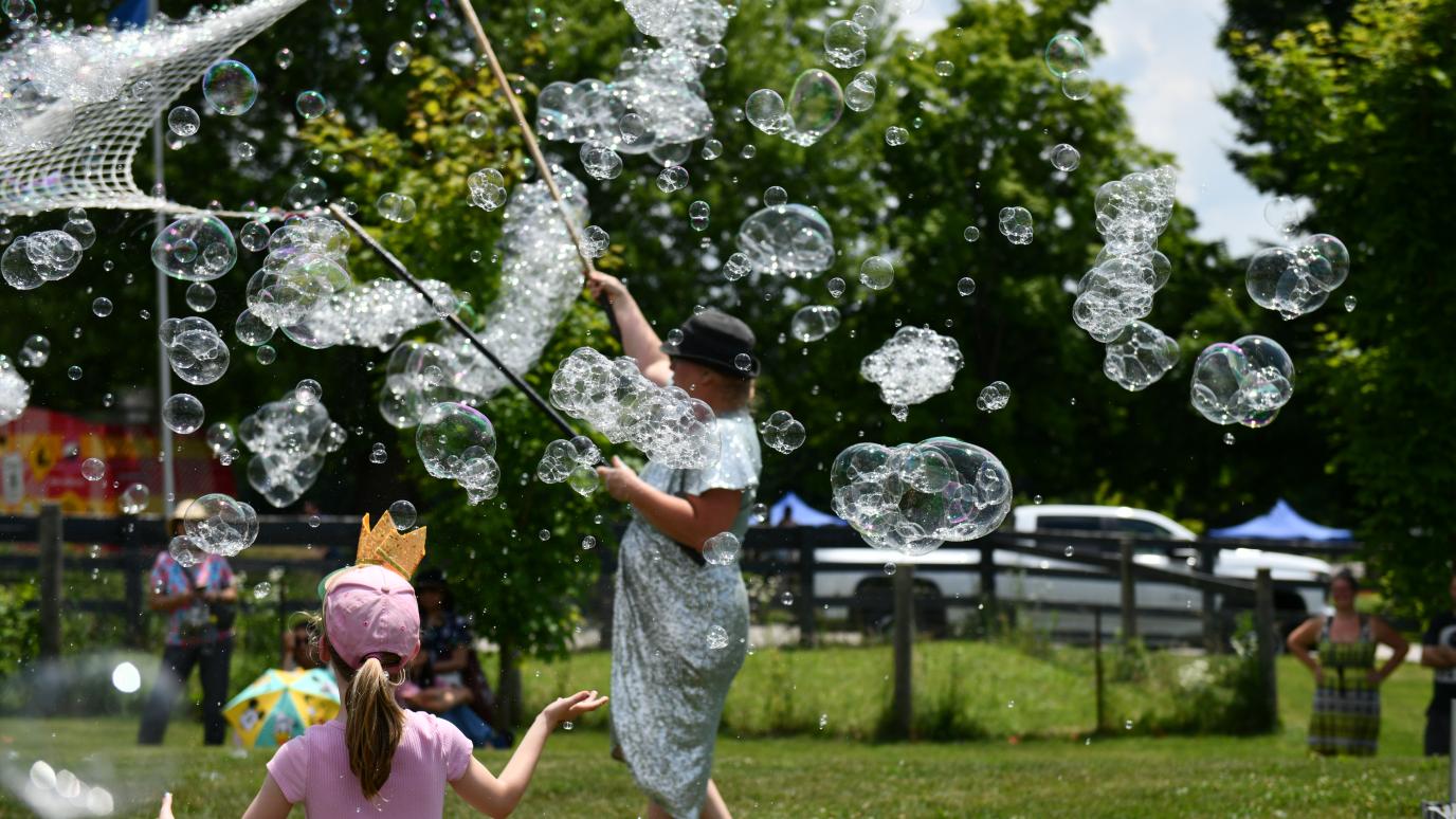 A performer at the 2025 Multicultural Festival holds two large sticks, creating large bubbles that children run to pop.