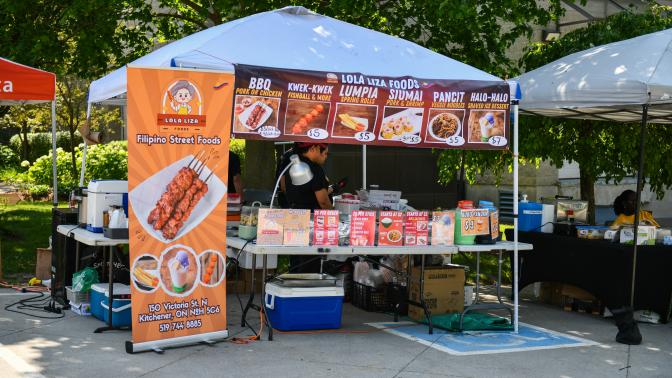 An outdoor food stall for "Lola Liza Foods" features banners advertising Filipino street foods like BBQ, lumpia, and halo-halo under a white canopy tent.