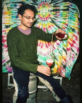 Person in a green sweater sits on a keg, pouring a drink dramatically between two glasses in front of a colourful tie-dye backdrop.