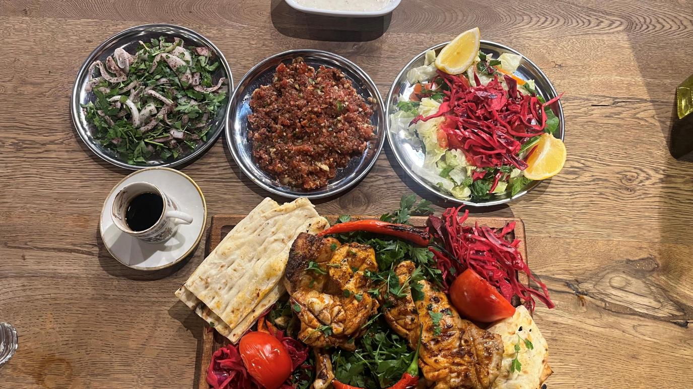 A Turkish grilled chicken meal served on a wooden table, featuring a large platter of grilled chicken with flatbread, parsley, grilled tomato and pepper, and red cabbage. Accompanying side dishes include a salad with lemon wedges, a minced vegetable and bulgur salad (possibly kısır), a fresh onion and parsley salad, a yogurt-based dip or sauce, and a cup of Turkish coffee