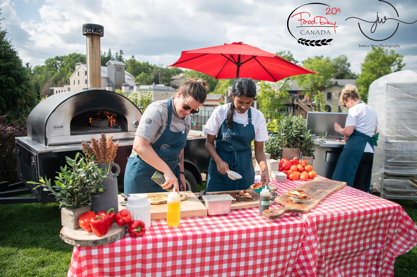 Two individuals prepare food on a table covered in a red gingham cloth. Behind them is an outdoor oven, and in the distance is the Elora Mill. Another person prepares more food on a barbeque beside them.